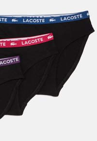 Trīs melni apakšveļas bikses ar kontrastējošiem jostas saites: zila, sarkana un violeta, visi ar Lacoste logotipu un gludu, kokvilnas tekstūru.