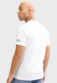 Hvid bomulds T-shirt med korte ærmer og en rund halsudskæring. Har teksten "REBEL" på venstre ærme. Bæres med denim jeans.