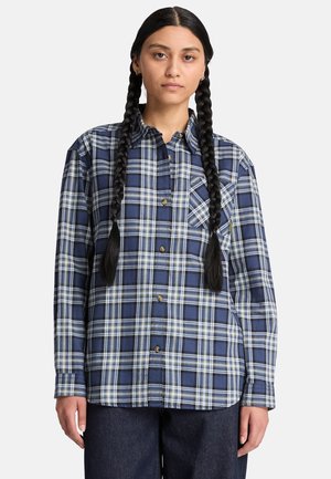 Femme aux longs cheveux tressés portant une chemise à carreaux bleue et blanche boutonnée et un pantalon foncé, debout devant un fond uni.