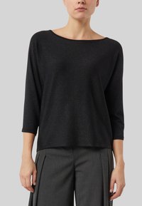 Czarna bluzka z długim rękawem o luźnym kroju i szerokim dekolcie, z delikatnym połyskiem. W połączeniu z plisowanymi szarymi spodniami.