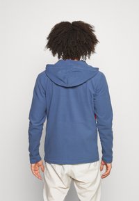 Felpa con cappuccio blu caratterizzata da una texture liscia, maniche lunghe e un dettaglio centrale a cucitura. La vista posteriore mette in risalto il design a vestibilità rilassata.