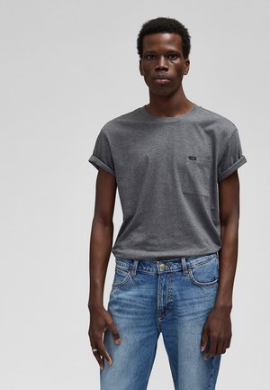 Młody mężczyzna ubrany w szarą koszulkę z podwiniętymi rękawami i kieszonką na piersi oraz niebieskie dżinsy, stojący na tle jednolitego szarego tła.