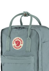 Sac à dos en tissu gris avec deux poignées supérieures, présentant un patch logo rond et une poche zippée sur le devant. Texture lisse avec des coutures renforcées.