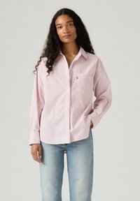 Camicia fucsia chiaro con righe verticali bianche, dotata di taschino frontale e maniche lunghe, abbinata a jeans blu chiaro.
