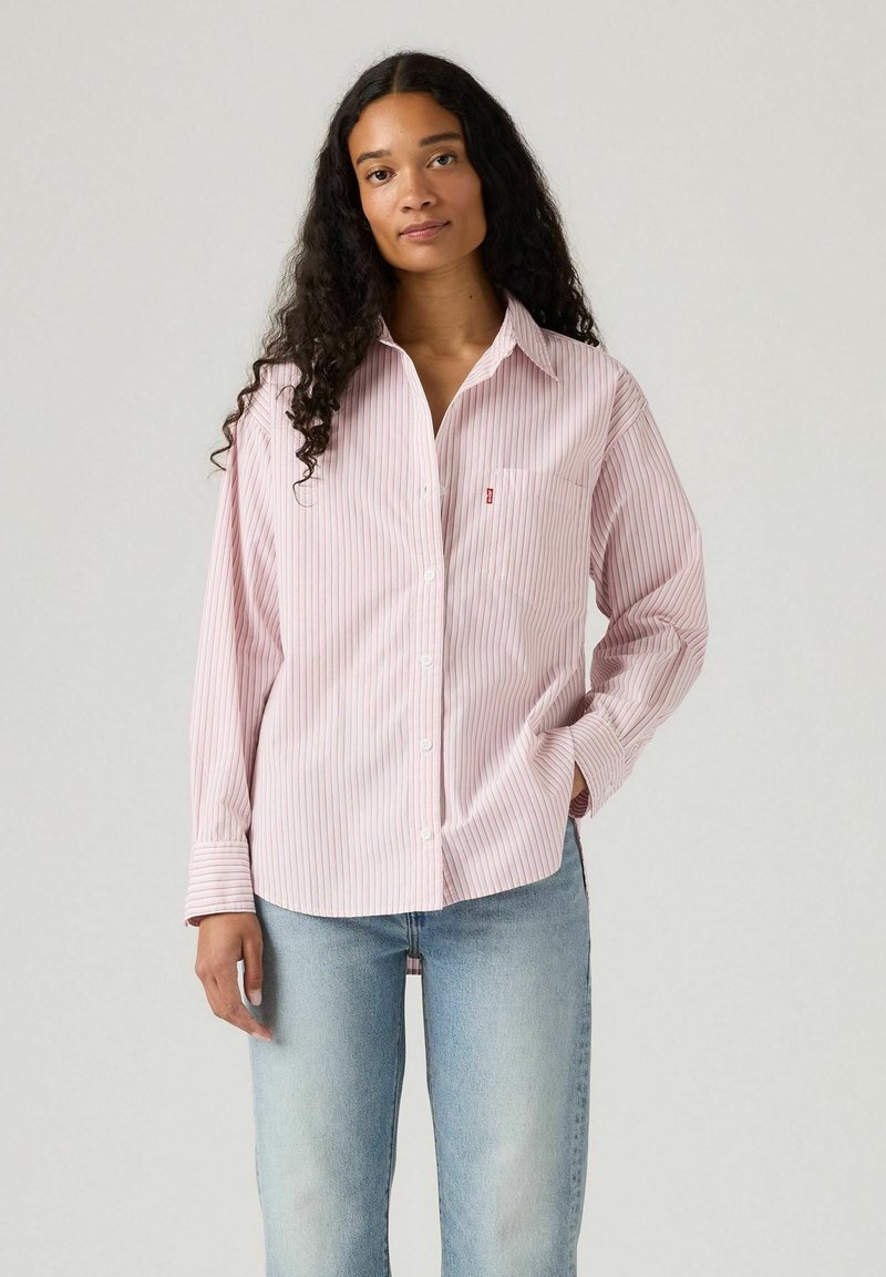 Camicia fucsia chiaro con righe verticali bianche, dotata di taschino frontale e maniche lunghe, abbinata a jeans blu chiaro.