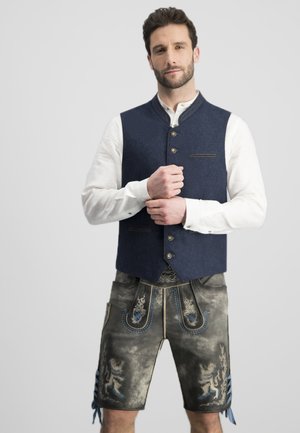 Marineblaue Wollweste mit hohem Kragen und vorderem Knopfverschluss, kombiniert mit grauen Shorts, die mit blauen bestickten Mustern und dekorativen seitlichen Fransen verziert sind.