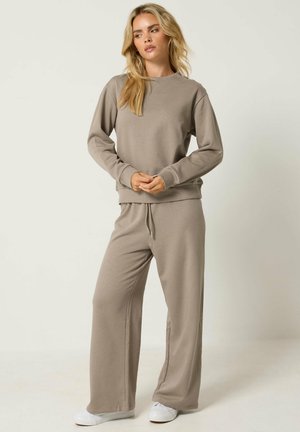 Frau steht, trägt einen langärmeligen taupefarbenen Sweatshirt und weitgeschnittene Hosen mit weißen Sneakern vor einem schlichten hellen Hintergrund.