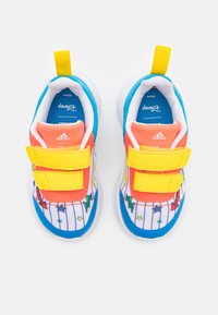 Zapatillas infantiles con correas de velcro amarillas, acentos azules y naranjas, suelas blancas y patrones coloridos de estrellas y rayos sobre un fondo blanco.