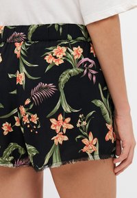 Shorts noirs à motif floral éclatant en orange et vert, avec un ourlet effiloché. Une taille élastique et des fentes sur les côtés sont visibles.