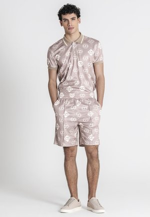 Ung mand i beige matchende mønstret poloshirt og shorts med hænderne i lommerne, iført beige sneakers, stående mod hvid baggrund.