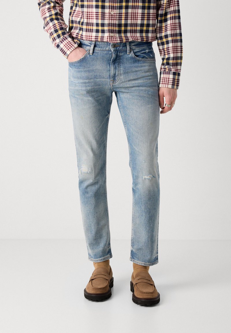 Jeans in denim blu chiaro con vestibilità slim, caratterizzati da dettagli strappati e una leggera sbiaditura. Indossati con una camicia a quadri e mocassini marroni.