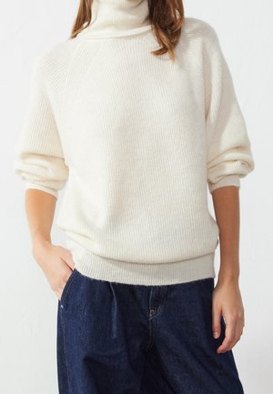Personne portant un pull à col roulé côtelé crème avec les manches retroussées et un jean denim bleu foncé, main dans la poche, debout devant un fond blanc.