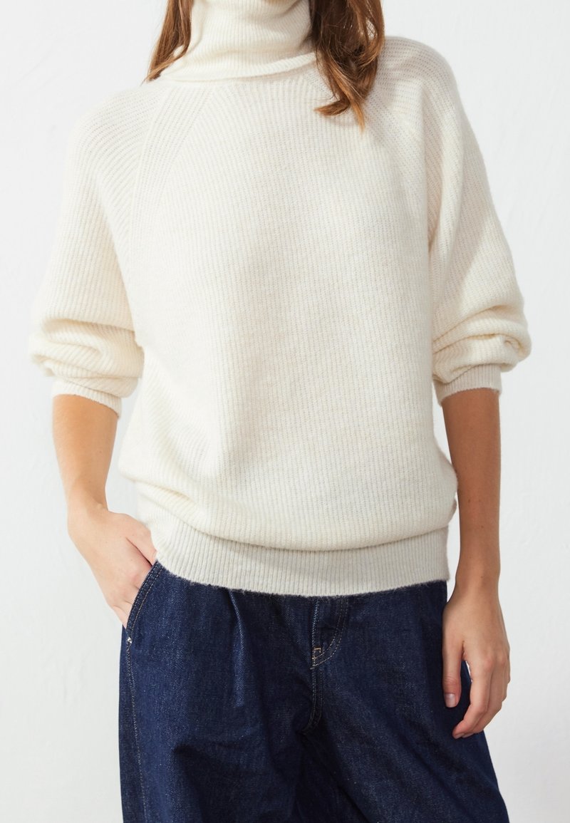 Personne portant un pull à col roulé côtelé crème avec les manches retroussées et un jean denim bleu foncé, main dans la poche, debout devant un fond blanc.