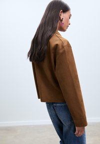 Veste en daim marron raccourcie avec des manches longues, présentant une coupe droite et un design minimaliste. Le modèle porte un jean bleu et des boucles d'oreilles remarquables.