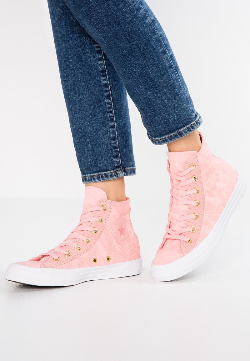 Pieds portant des baskets montantes roses avec des semelles blanches et des œillets dorés, assorties à un jean bleu retroussé sur un sol blanc.