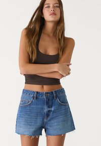 Farkkushortsit, joissa on revitty helma, haalistunut sininen pesu, viisi taskua ja etupuolella nappikiinnitys, yhdistettynä ruskeaan crop top -paitaan.