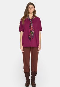 Luźna fioletowa bluzka z krótkimi rękawami, w zestawie z brązowymi spodniami o zwężanym kroju. Na szyi związany jest wzorzysty czarny szal z różowymi akcentami.