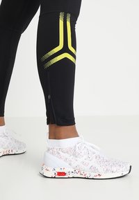 Czarne legginsy sportowe z żółtym ukośnym paskiem oraz zamek błyskawiczny na kostkach, zestawione z wielokolorowymi butami do biegania o wzorze.