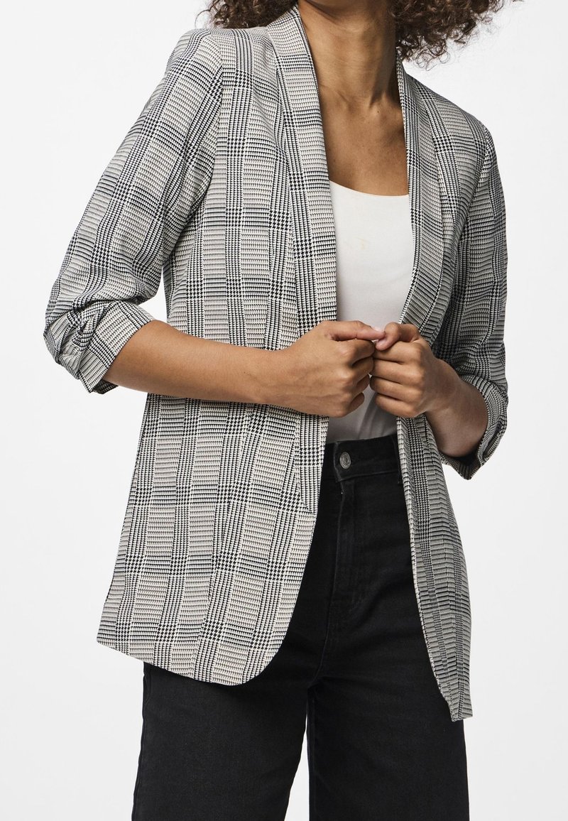Femme portant un blazer à carreaux noirs et blancs par-dessus un haut blanc et un pantalon noir, ajustant le devant du blazer avec les deux mains.