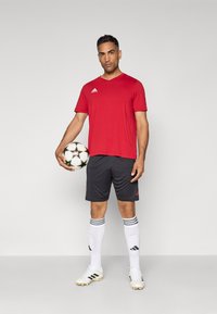 Maillot de sport rouge, shorts noirs, chaussettes rayées blanches et chaussures de football. Tenant un ballon de soccer à motifs, debout sur un fond neutre.