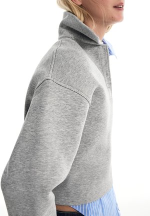 Femeie purtând un hanorac gri, cu mâneci lungi și guler înalt, peste o cămașă cu dungi albastre și albe, văzută din profil la nivelul taliei.