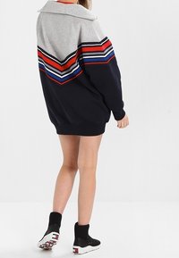 Persona vistiendo una sudadera grande con parte superior gris, parte inferior azul marino y franjas de chevron en rojo, blanco y azul, combinada con zapatillas tipo calcetín negras.