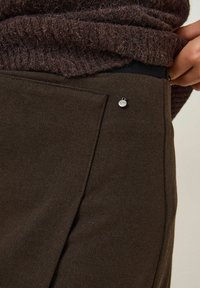 Gros plan sur la main d'une personne ajustant un pantalon marron foncé avec une poche arrière et un petit pin en métal rond près de la taille.