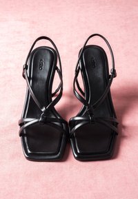 Czarne sandały na cienkich paskach z kwadratowym noskiem i paskiem z tyłu, na różowej teksturowanej powierzchni.