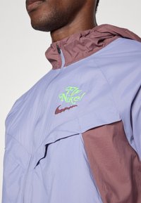 Άνδρας φορώντας ένα ανοιχτό μωβ και καφέ αδιάβροχο μπουφάν Nike με πράσινο λογότυπο "Fly Nike!" και καφέ swoosh στο στήθος, κλειστό.