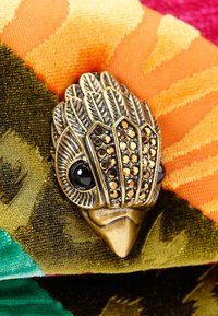 Spilla in bronzo a forma di uccello con accenti gioiello, posata su un tessuto vivace nei toni dell'arancione, verde e rosa. Presenta una texture dettagliata simile a piume.