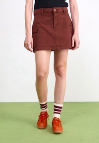 Gonna cargo in cotone marrone con tasche frontali a patta, chiusura a bottone e taglio dritto. Abbinata a sneaker arancioni e calzini a righe.