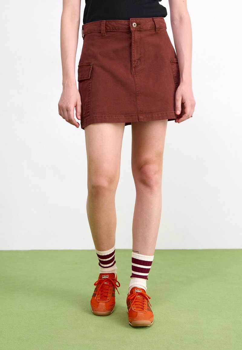 Gonna cargo in cotone marrone con tasche frontali a patta, chiusura a bottone e taglio dritto. Abbinata a sneaker arancioni e calzini a righe.