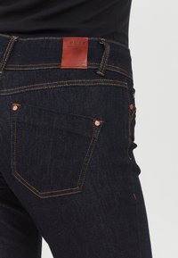 Vaqueros de mezclilla azul oscuro con costuras naranjas, que presentan un parche trasero de cuero etiquetado "PULZ" y detalles de hardware en color cobre.