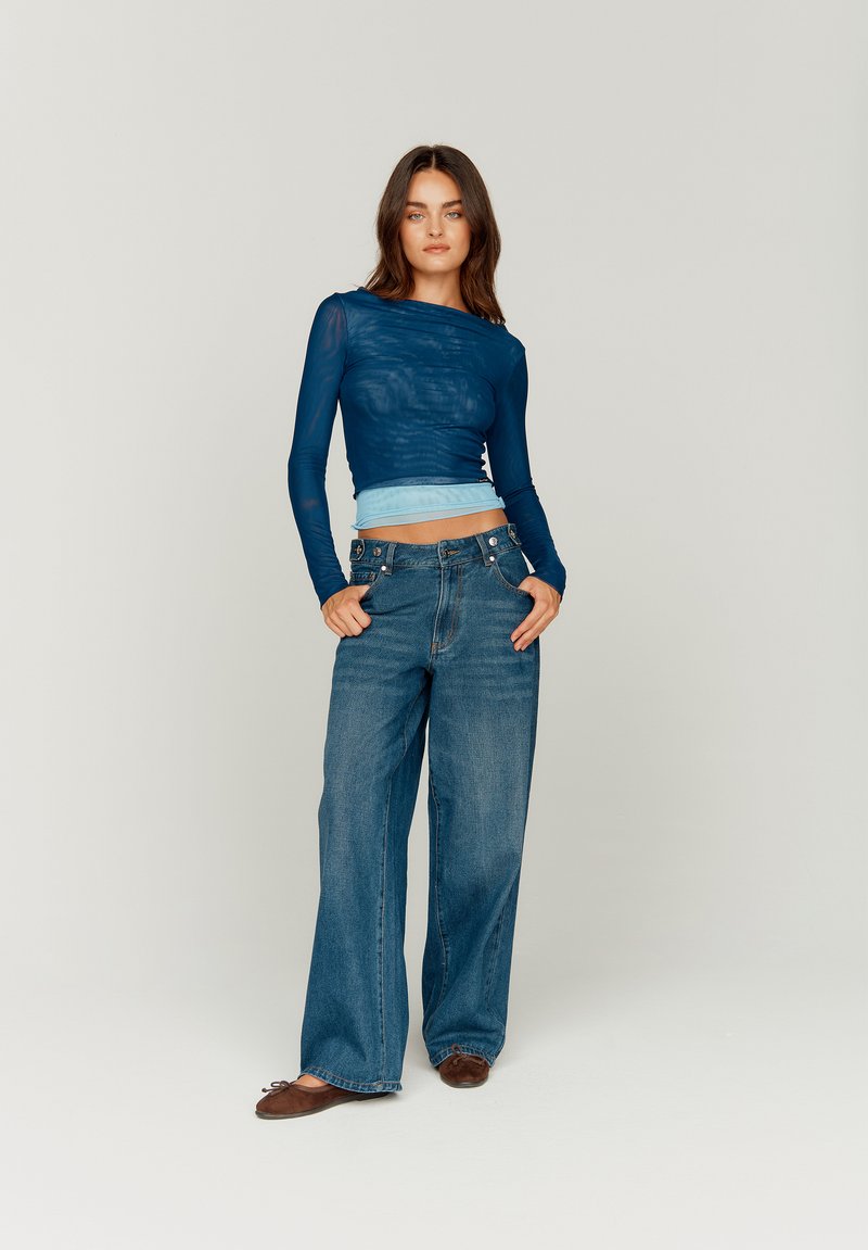 Long-sleeve, sheer dark blue top with a light blue waistband paired with wide-leg dark jeans. Brown flat shoes with a rounded toe.