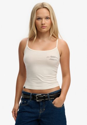 Mujer rubia con un top crema de tirantes finos que lleva el texto "Athletica 1985", jeans azules y un cinturón negro con tachuelas, de pie con una mano en el bolsillo.