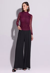 Bordowa bluzka z długim rękawem z marszczonym materiałem i kwiatowym akcentem. Czarne spodnie o szerokich nogawkach. Modelka ma na sobie czarne, futerkowe sandały.