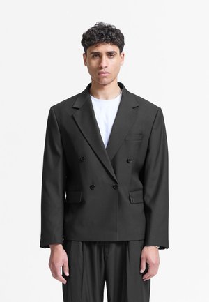 Mężczyzna z krótkimi kręconymi włosami nosi czarną dwurzędową marynarkę na białej koszuli, stojąc na tle jednolitego białego tła.