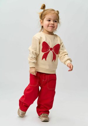 Beige sweatshirt with a large red bow graphic, paired with loose red pants. Textured gold shoes complete the outfit. Soft fabric, casual fit.