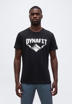 Férfi áll szemben, fekete Dynafit pólót visel hegyi logóval és kék Dynafit nadrágot egyszínű fehér háttér előtt.