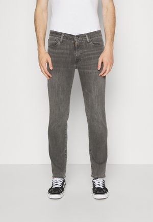 Homme portant un jean gris slim, un débardeur blanc et des baskets noires avec des lacets blancs, debout devant un fond blanc uni.