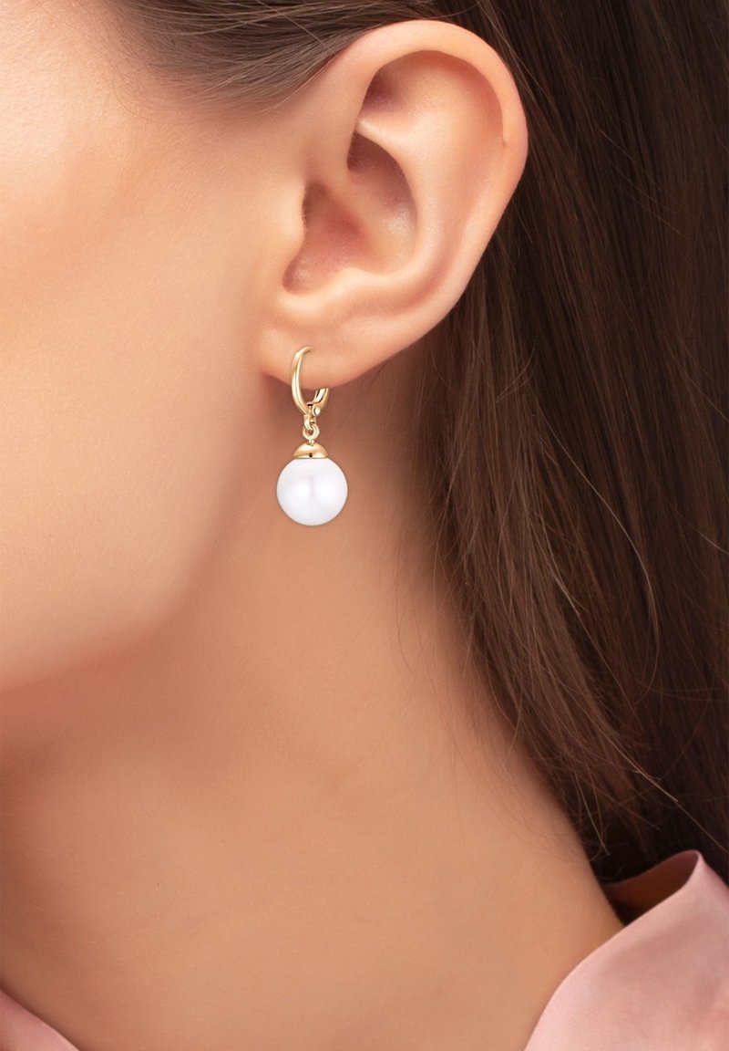 Gros plan sur l’oreille d’une femme portant une boucle d’oreille créole en or avec un pendentif en perle blanche.
