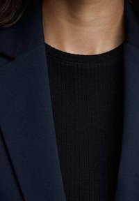 Tissu de blazer bleu marine à la texture lisse, superposé à un haut en maille côtelée noire. Le col et l'encolure sont visibles, mettant en valeur des lignes épurées.