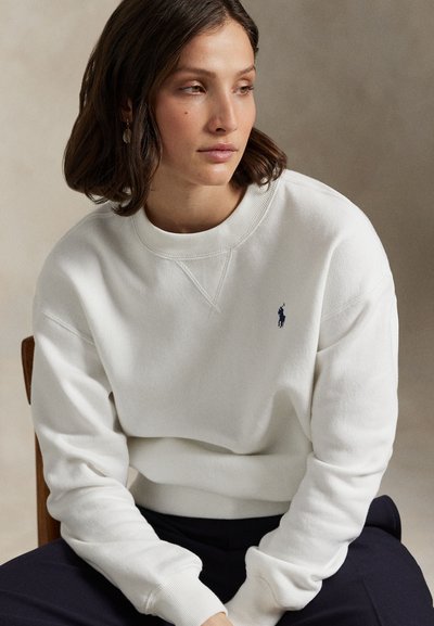 Γυναίκα με καστανά μαλλιά μέχρι τους ώμους, που φοράει λευκό φούτερ με μικρό σκούρο λογότυπο, καθισμένη μπροστά σε ουδέτερο φόντο.