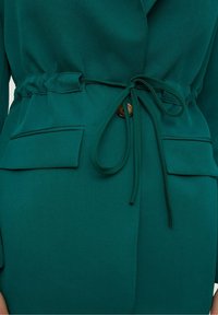 Vihreä kietaisupaita, jossa on vyötärölle kiinnitettävä vyö, sileä rakenne ja kaksi läpällistä taskua. Yksityiskohtia ovat kankaasta tehty nauha ja pyöreä nappi korostuksena.