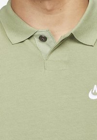 Jasnozielona koszulka polo z kołnierzykiem, z jednym zapiętym czarnym guzikiem i częściowym białym logo Nike na piersi.