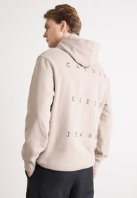 Beige kapucnis pulóver, a hátán függőlegesen valósidejű felirattal: "CALVIN KLEIN JEANS" fekete színben. Pamut keverék, puha tapintású, laza fazon.