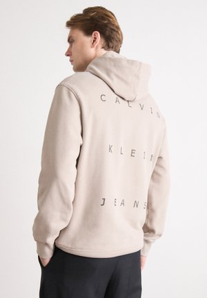 Felpa beige con cappuccio, caratterizzata dalla scritta "CALVIN KLEIN JEANS" stampata verticalmente sul retro in nero. Tessuto misto cotone, morbido al tatto, vestibilità rilassata.