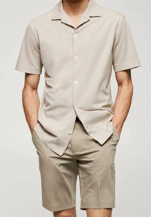 Homme portant une chemise beige à manches courtes boutonnée et un short beige assorti arrivant aux genoux, les mains dans les poches.