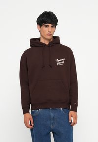 Bruine hoodie van een katoenmix met een voorkantzak, capuchon met trekkoord en een wit "Tommy Jeans" logo aan de linkerkant. Casual fit ontwerp.