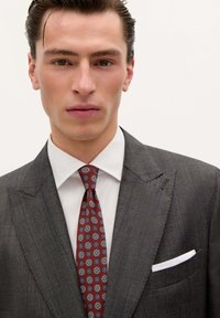 Traje en gris oscuro con finas rayas en espiga, camisa blanca y corbata burdeos con patrones circulares azules y amarillos. Pañuelo blanco en el bolsillo visible.